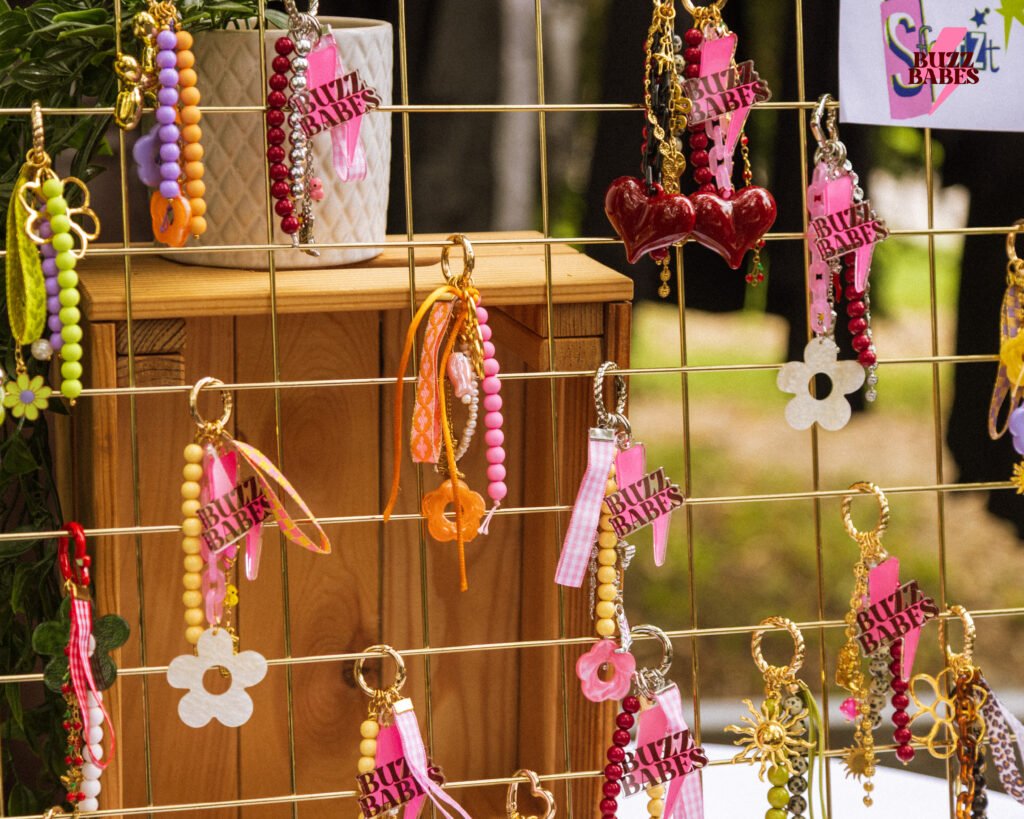 Handgemachte Buzz Babes Schlüsselanhänger und Schmuckstücke in bunten Farben, ausgestellt auf einem goldenen Gitter bei einem Marktstand.