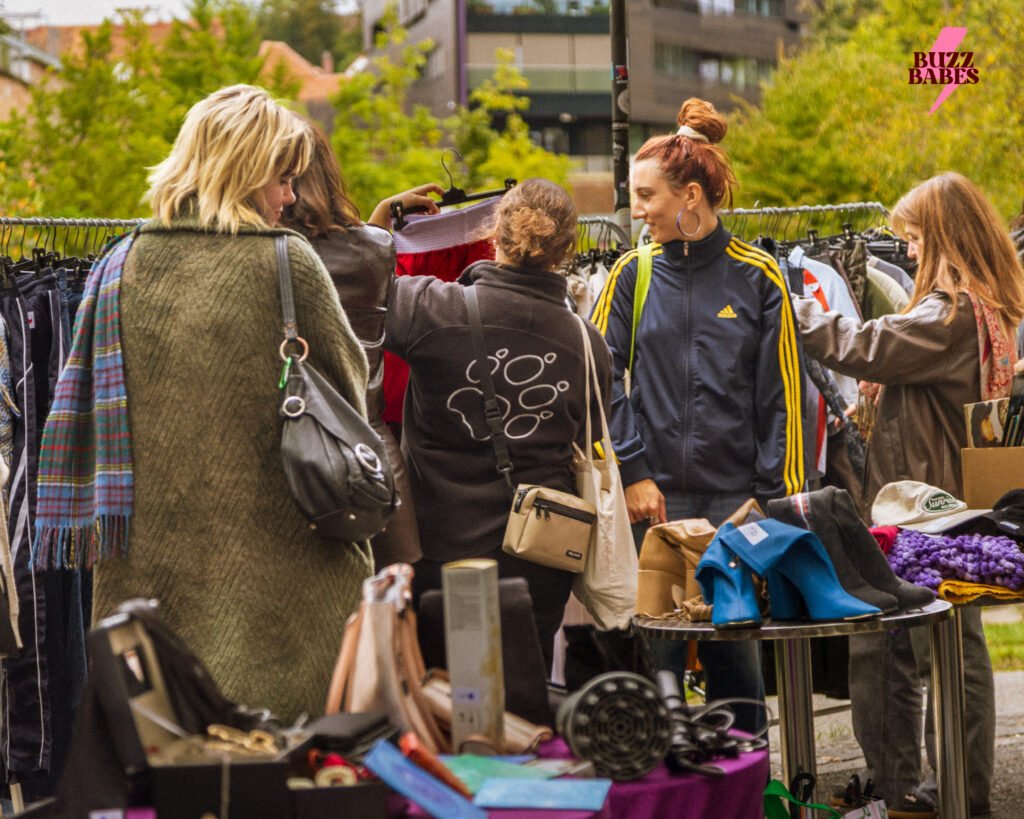 Besucherinnen stöbern an einem Buzz Babes Flea Market durch Kleidung und Accessoires an Kleiderstangen im Freien.