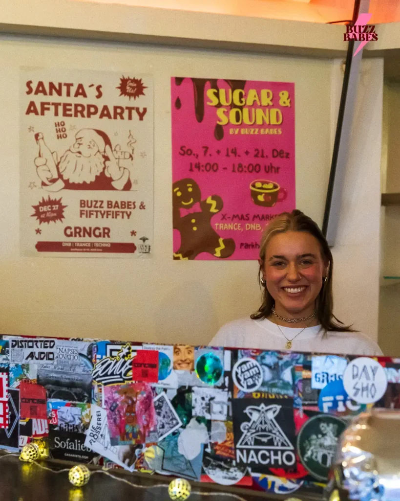 DJ hinter einem mit Stickern beklebten DJ-Pult bei einem Buzz Babes Event, im Hintergrund bunte Eventposter an der Wand.