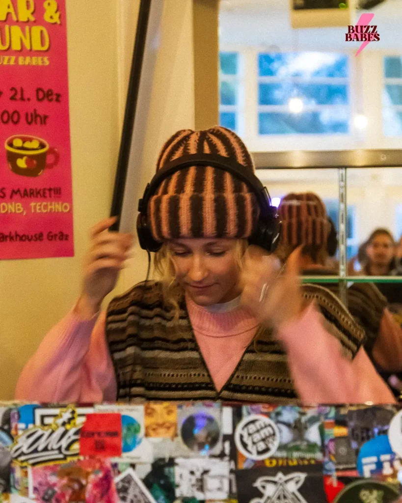 DJ mit Kopfhörern hinter einem mit Stickern beklebten DJ-Pult bei einem Buzz Babes Event.